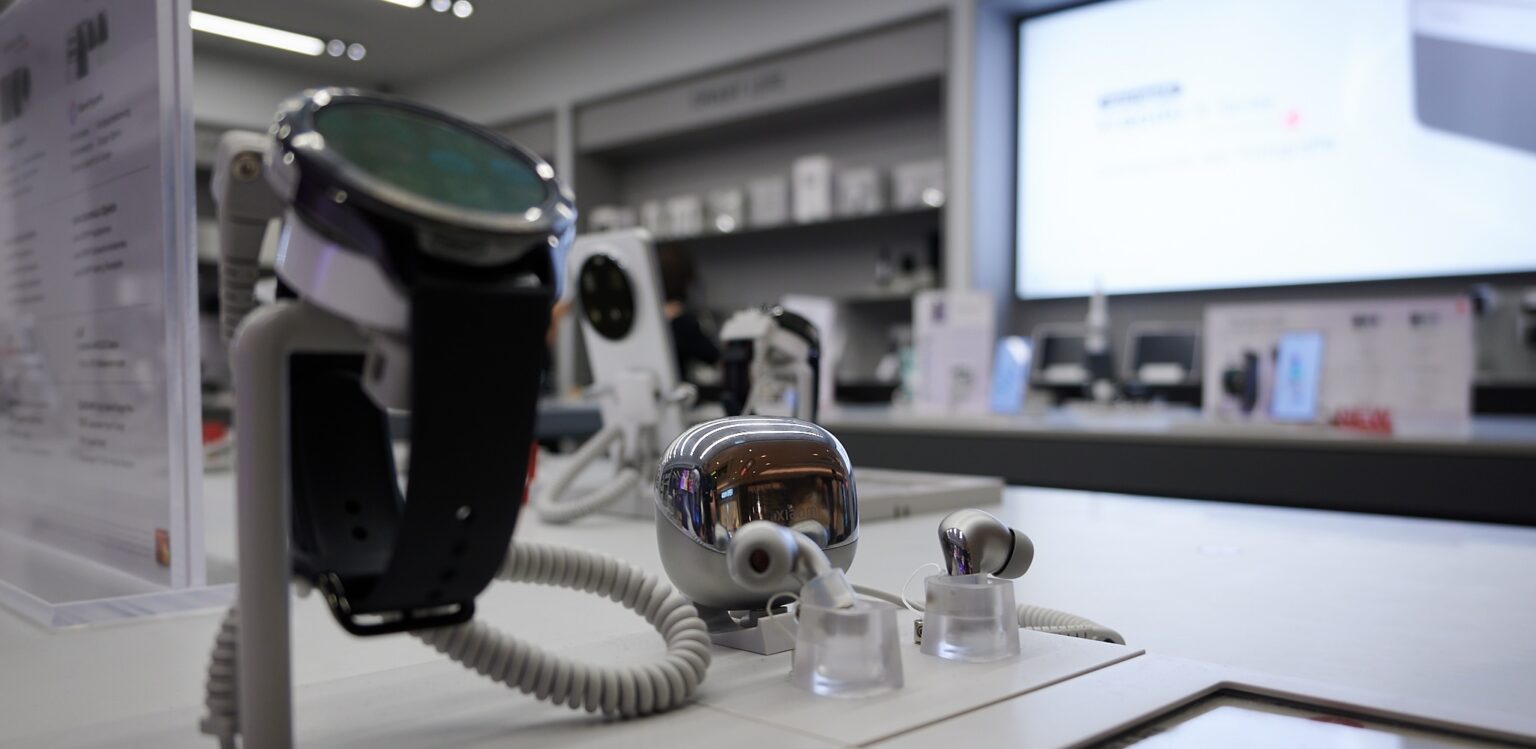 Blick in den Xiaomi-Store im Hintergrund mit Nahaufnahme auf eine Smartwatch und ein Ohrhörer-Case (freigestellt).