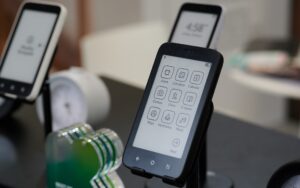 Freigestelltes Kompakt in Schwarz vor weiteren zwei Kompakt-Geräten im Hintergrund. Mit Kachel-Oberfläche für Apps sichtbar.