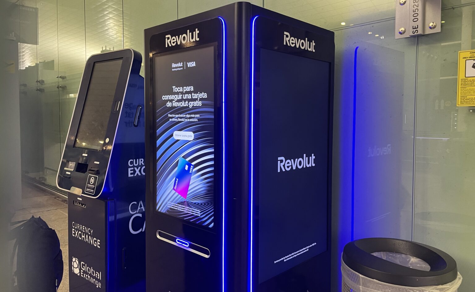 Ein Revolut-Automat mit zwei großen Displays zur Kartenausgabe.Daneben ein Mülleimer und ein Geldautomat.